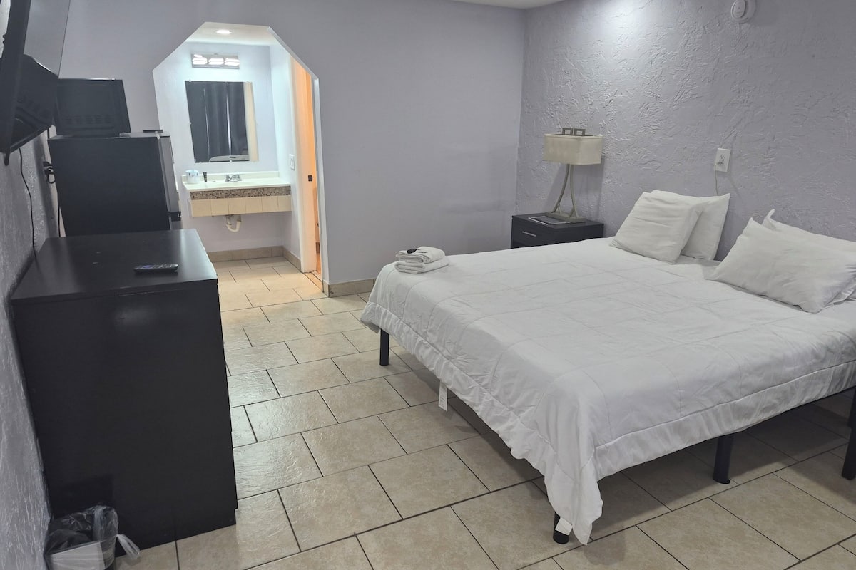 Una habitación de hotel con cama, mesita de noche, lámpara y vista al baño.