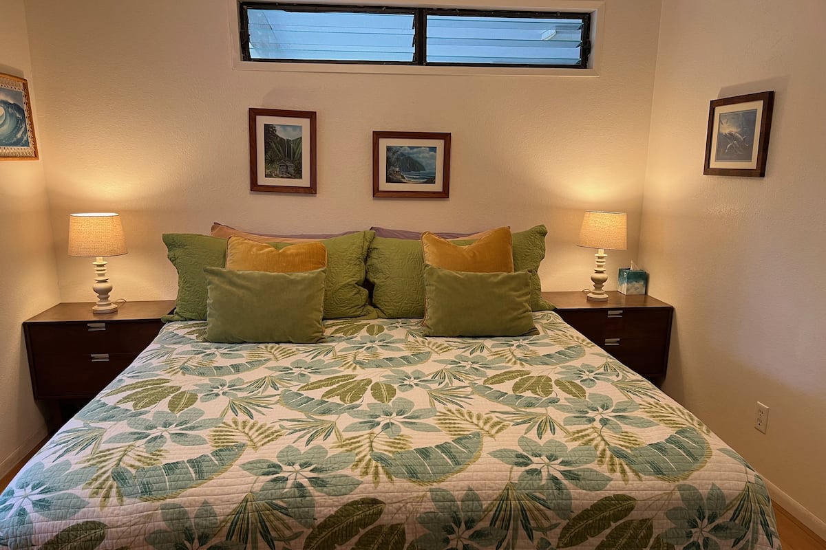 A bedroom with a bed, two nightstands, two wall-mounted lamps, a window, and two framed pictures on the wall.