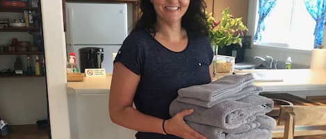 A woman holding a stack of towels in a kitchen with wooden cabinets, a coffee maker, and a window with curtains.