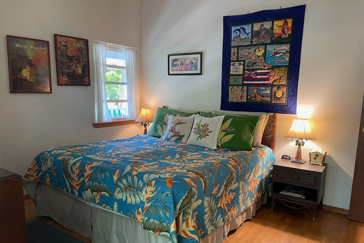 A bedroom with a floral bedspread, a quilt with various images, a nightstand with a lamp, and framed pictures on the wall.