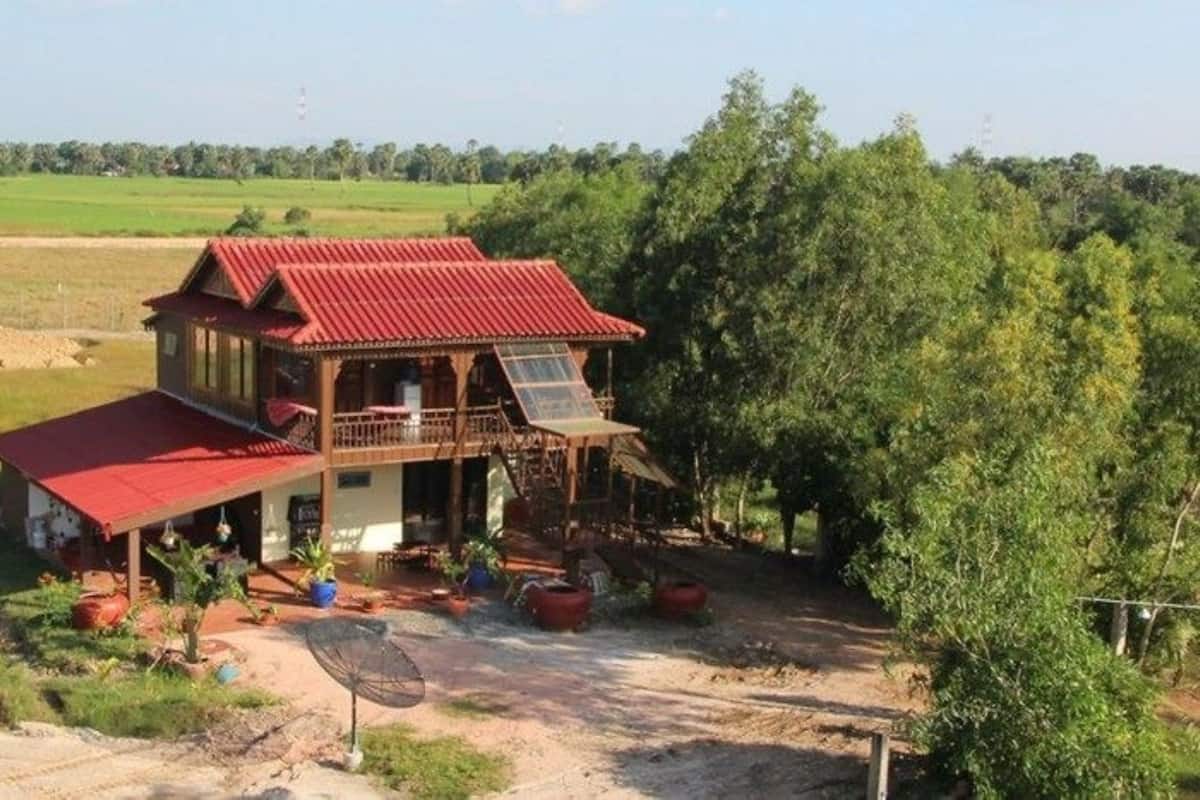A rustic wooden house with a red roof, surrounded by greenery and fields.
