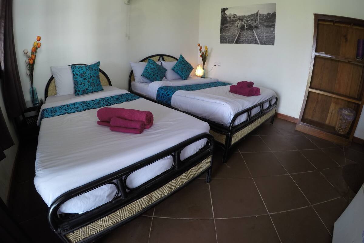A hotel room with two beds, each with white linens and teal patterned bedspreads. There is a wall-mounted fan above the beds, a framed photograph on the wall, and a wooden cabinet to the right.