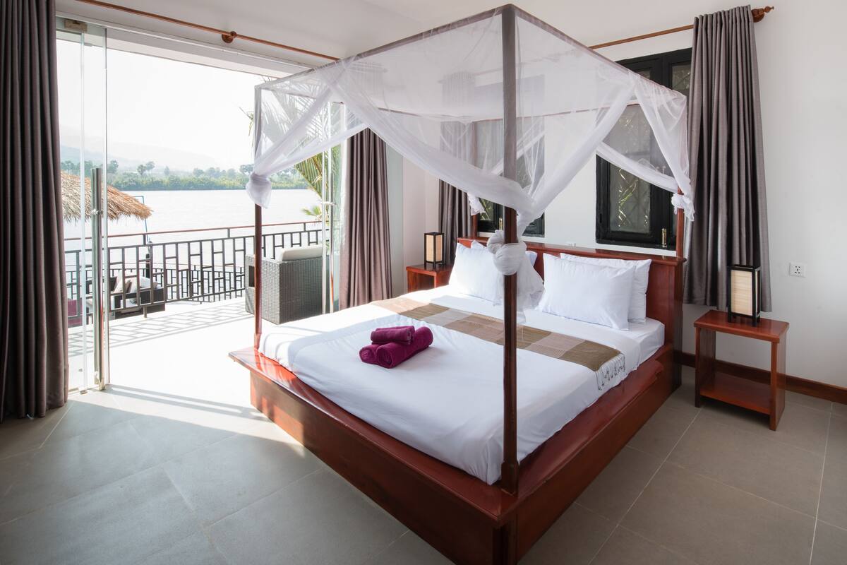 A four-poster bed with white linens and a canopy, a wooden nightstand, and a view of a balcony with a railing.