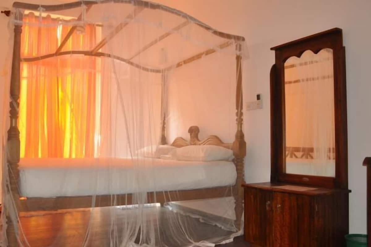 A room with a four-poster bed covered by a mosquito net, a wooden nightstand, and a mirror.