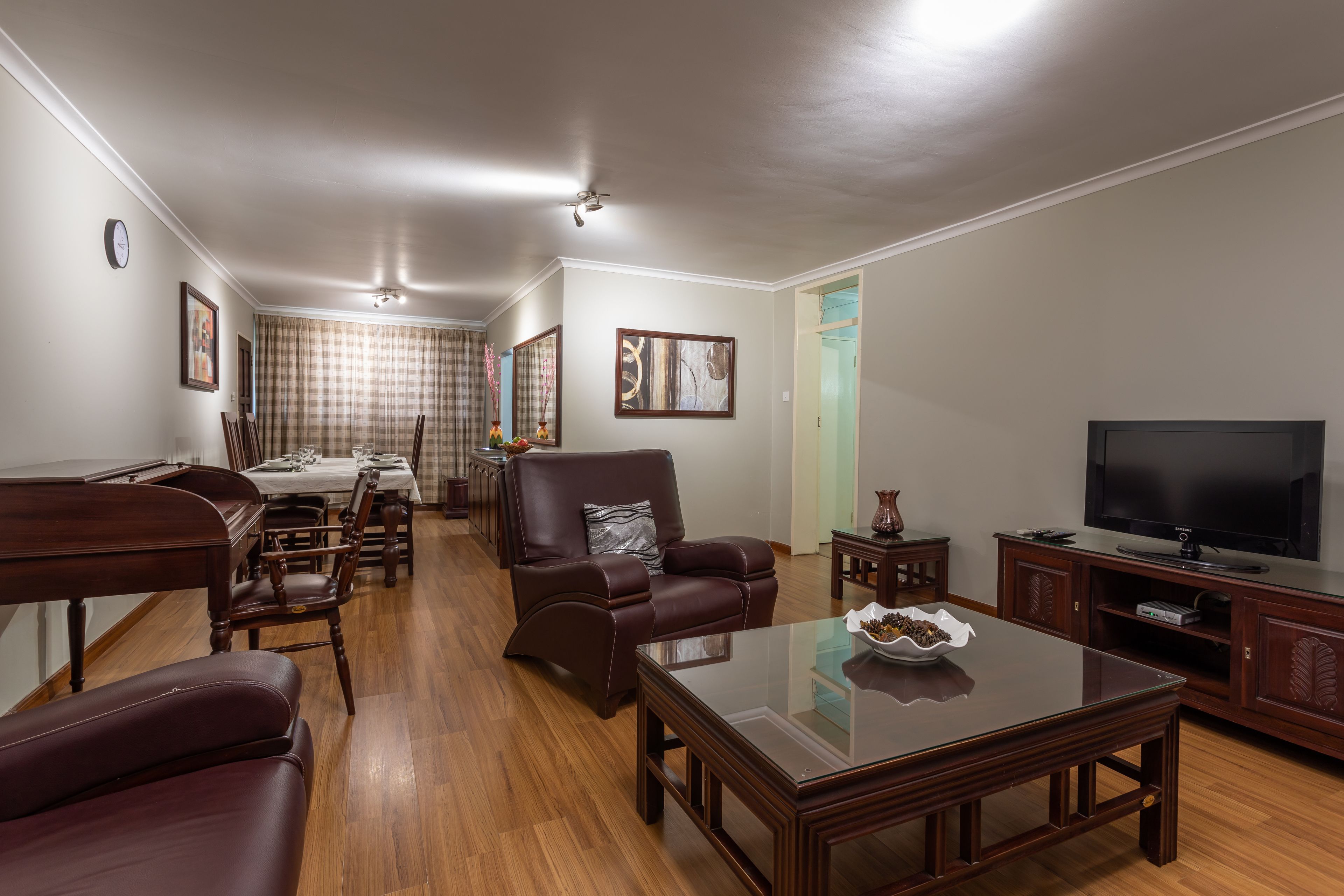 A spacious living room with a dining area, wooden furniture, a flat-screen TV, and a clock on the wall.
