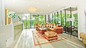 A spacious living room with a sectional sofa, a coffee table, and a red patterned rug. There is a reception desk with a vase of flowers and a large window offering a view of greenery outside.
