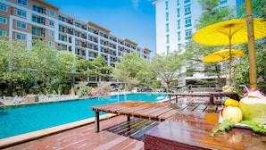 A swimming pool with a wooden deck and tables, surrounded by greenery and modern buildings.