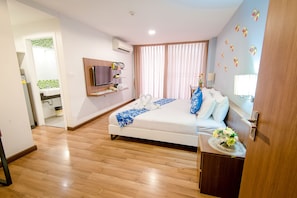 Une chambre d’hôtel moderne avec un plancher en bois, un lit avec une literie blanche, un téléviseur fixé au mur, une petite salle de bain avec une douche carrelée de carreaux verts et blancs, et une grande fenêtre avec des rideaux.