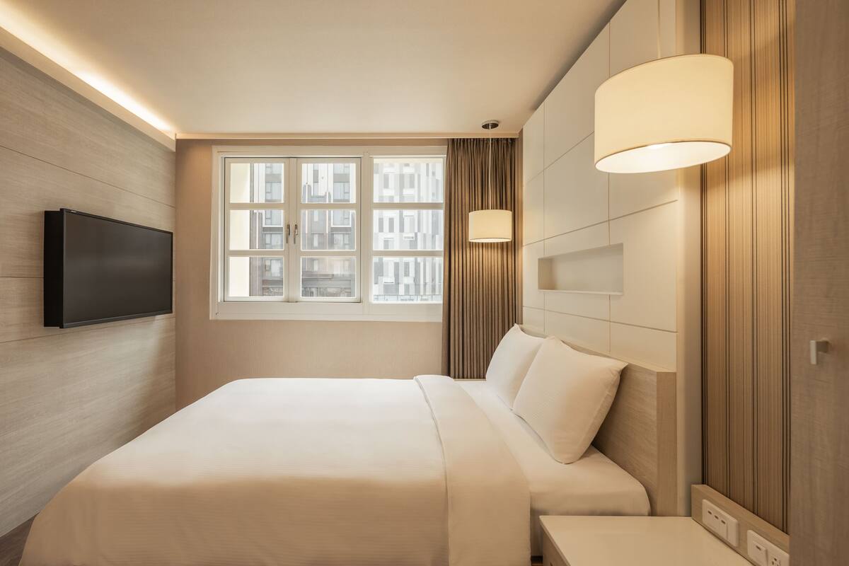 A modern hotel room with a large bed, a flat-screen TV mounted on the wall, and a window with curtains.