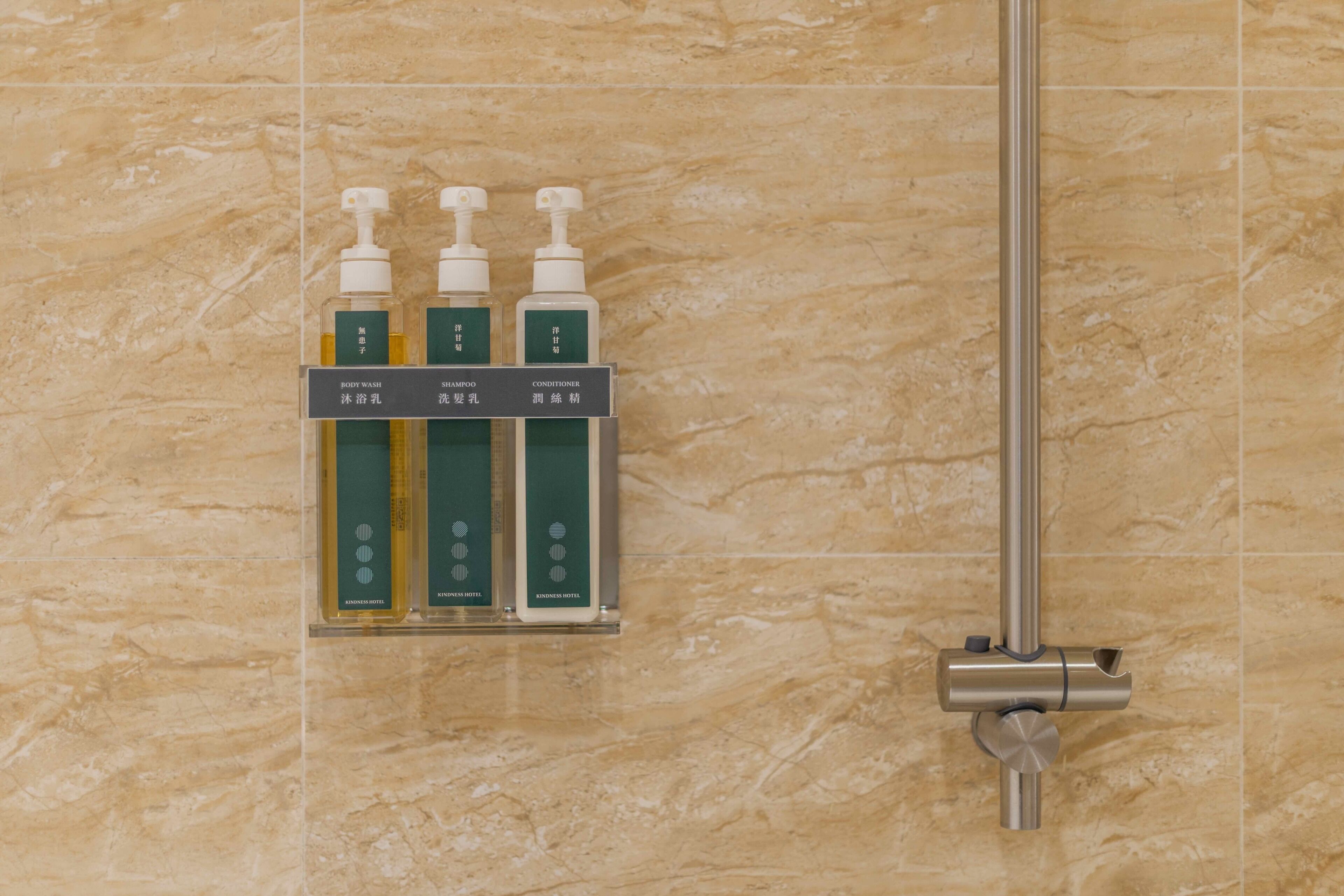 A bathroom with a marble wall, a shelf holding three bottles of body wash, shampoo, and conditioner, and a showerhead.