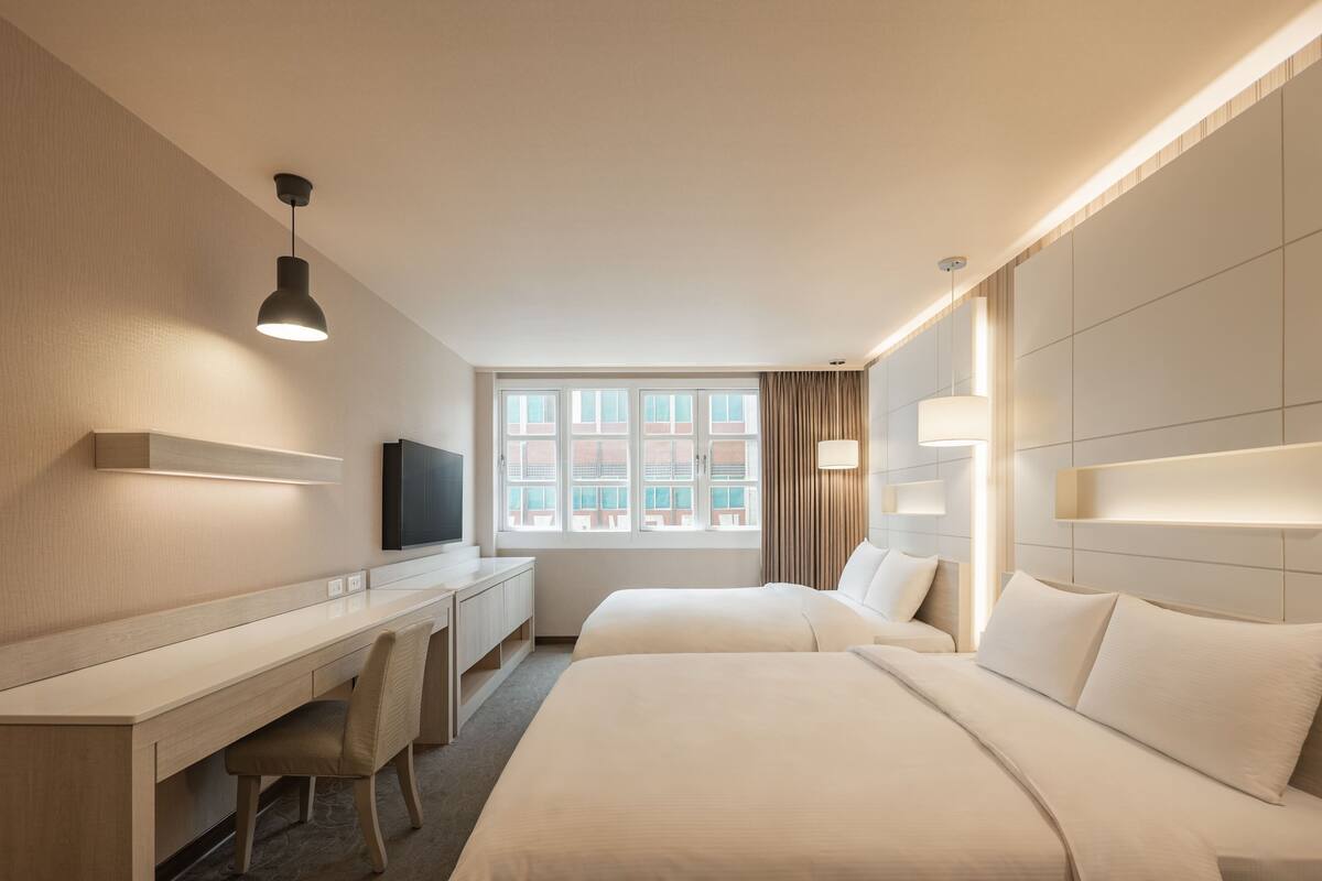 A hotel room with a desk, two beds, a television, and a large window with curtains.
