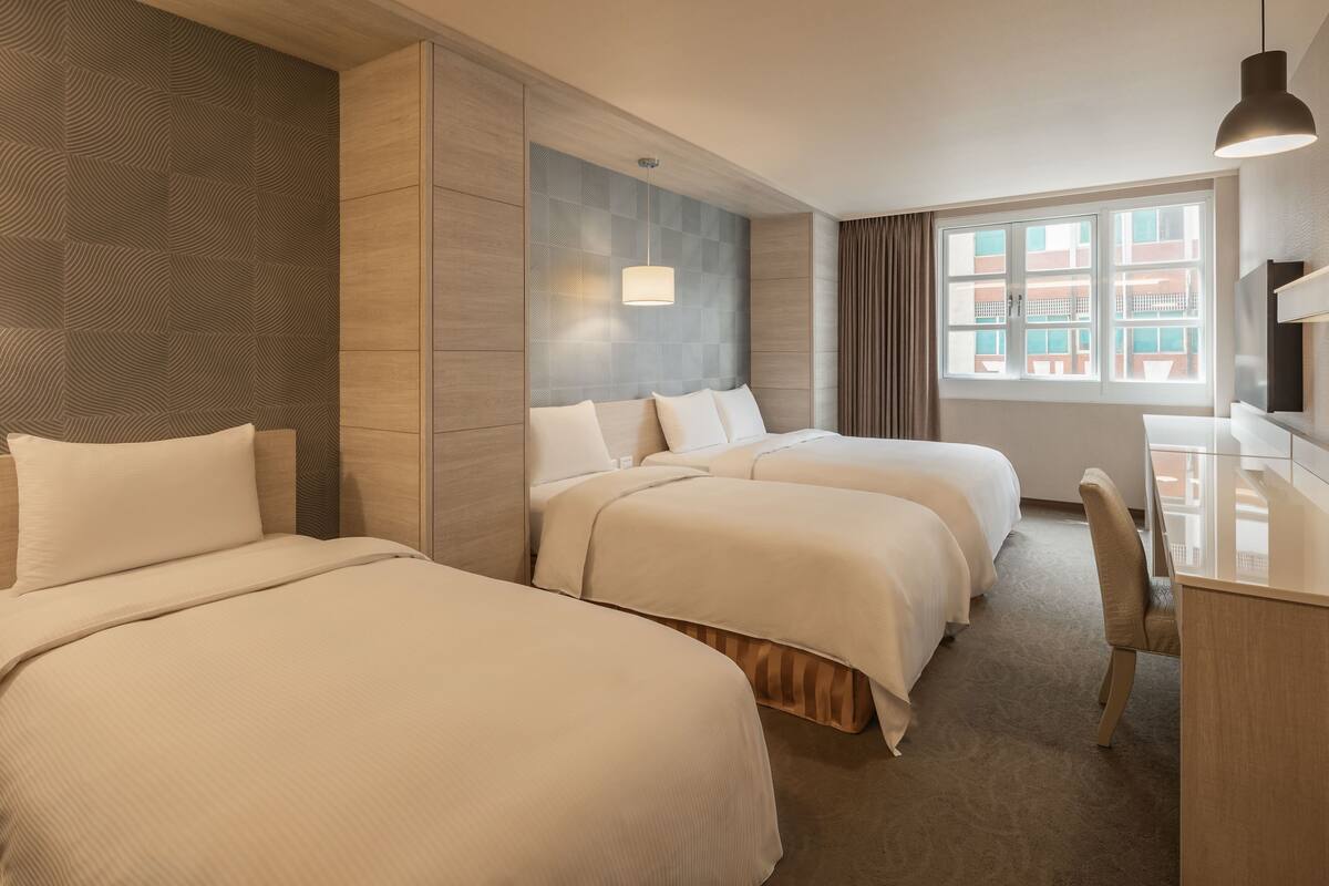 A hotel room with three beds, a desk, a chair, and a large window with a view of buildings.