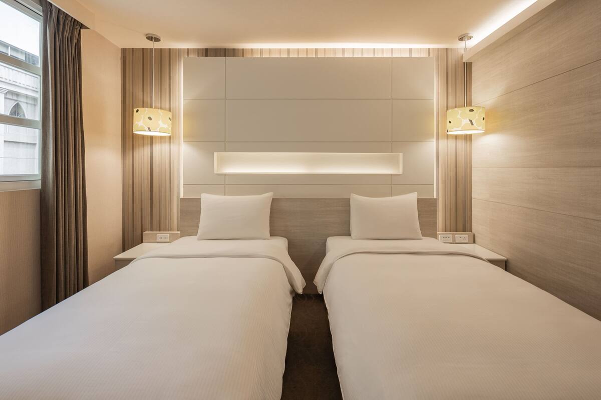 A hotel room with two single beds, a bedside table with a lamp, and a headboard with built-in lighting.