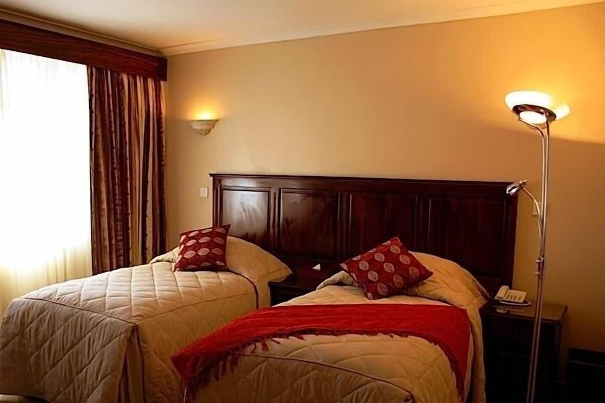 A hotel room with two beds, a wooden headboard, a bedside lamp, and a window with curtains.