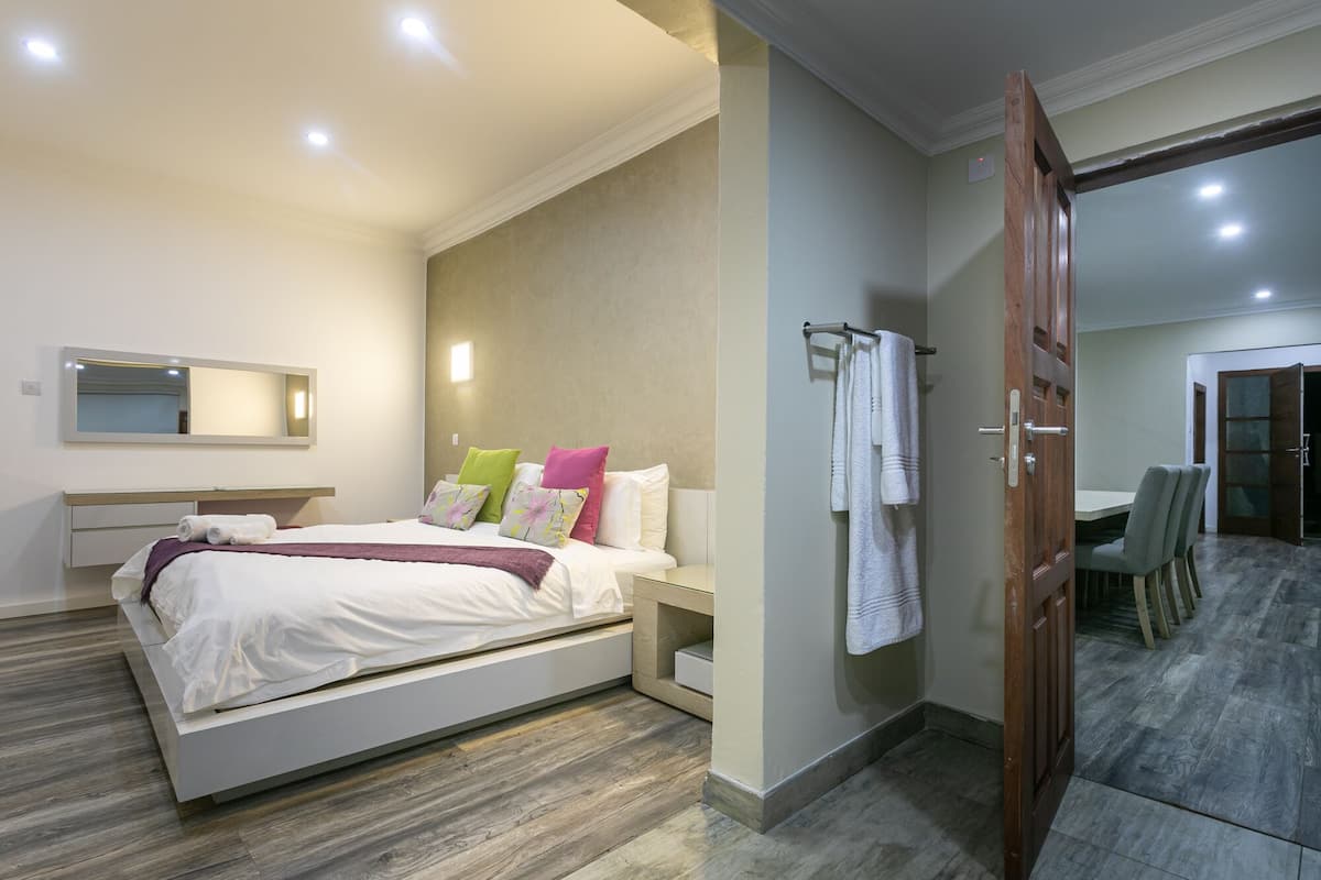 A modern bedroom with a bed, bedside table, and a mirror. There is a bathroom visible with a shower and a towel rack.