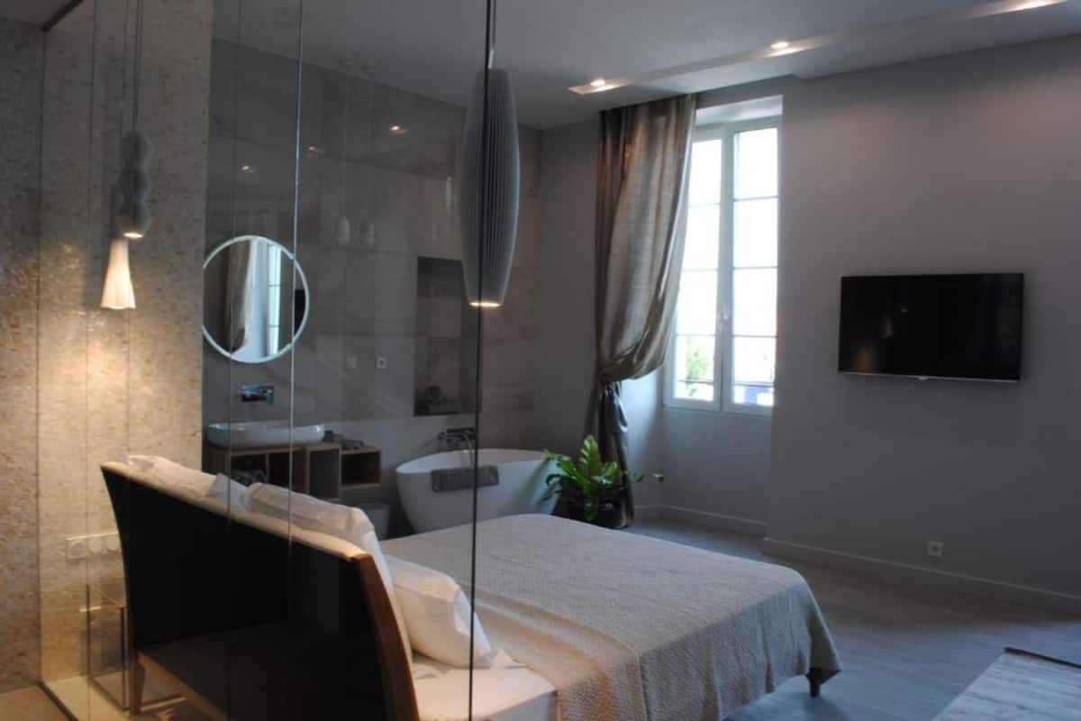 A modern bedroom with a bed, a large mirror, a bathtub, a TV, and a window with curtains.