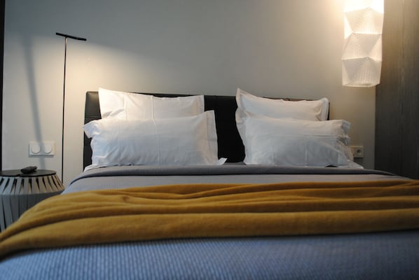 Una cama bien hecha con almohadillas blancas y una manta color mostaza, una mesita de noche con una lámpara, y una lámpara de pared.