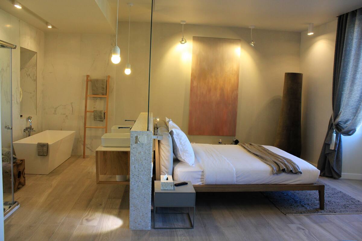 A modern bedroom with a large bed, bedside table, and a built-in desk with a chair. There is a bathtub and a shower in the adjacent bathroom.