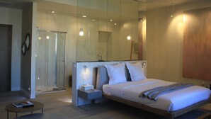 A modern bedroom with a large bed, a bedside table, a nightstand, a glass shower enclosure, and a small round mirror. - Les Suites Massena (Nice)