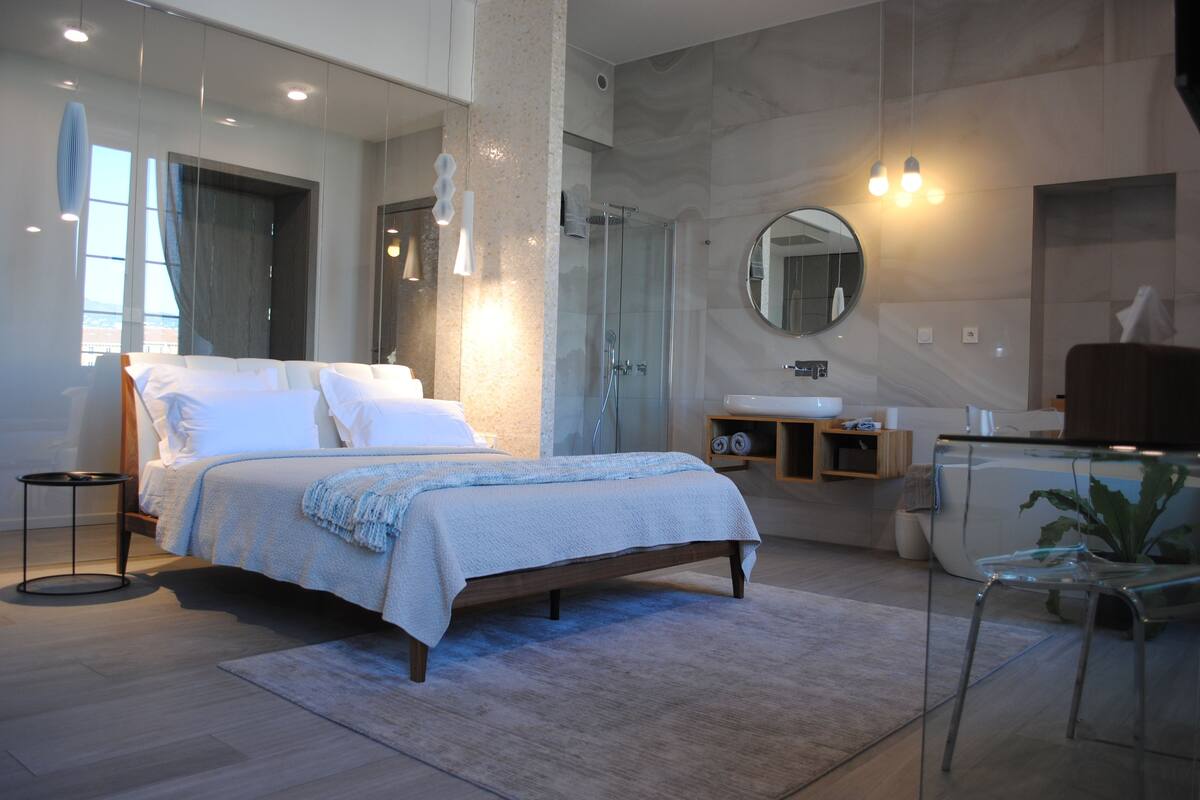 A modern bedroom with a large bed, a glass shower, and a round mirror above the sink.