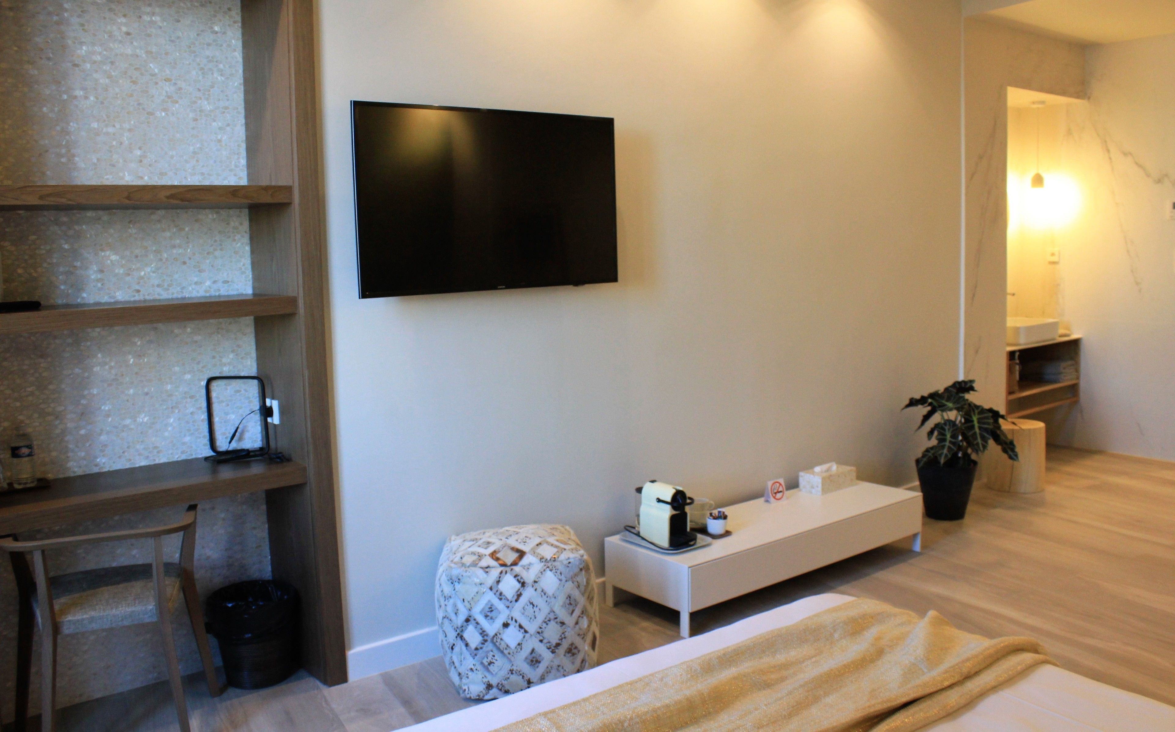 Habitación de hotel con cama, mesita de noche, televisor montado en la pared, pequeña mesita con lámpara, planta en maceta y espejo.