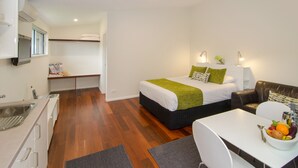 Habitación de hotel compacta con kitchenette, cama con ropa de cama verde, zona de comedor con frutero y sofá de cuero.