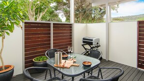 Un patio al aire libre con una mesa de vidrio, sillas de mimbre negro, una parrilla y plantas en macetas.