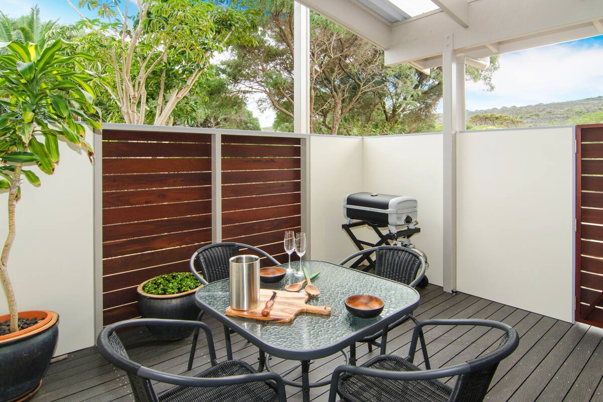 Una terrazza esterna con tavolo in vetro, sedie in vimini nero, un barbecue e piante in vaso.