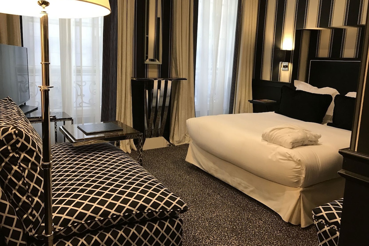 A hotel room with a bed, a desk, a chair, a lamp, and curtains.