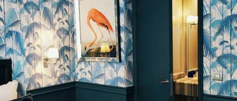 Hotellihuone, jossa on sänky, yöpöytä, kehystetty flamingoaiheinen kuva ja ovi, joka johtaa kylpyhuoneeseen.