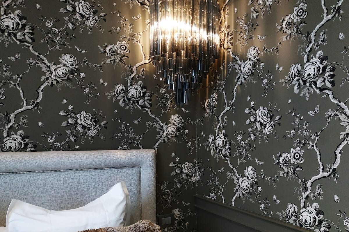 A bedroom with a dark floral wallpaper, a modern chandelier, a grey headboard, and a small wooden stool.