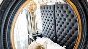 Un dormitorio con cabecera capitonada, una cama con sábanas blancas y dos almohadas, una mesita de noche con lámpara y un espejo con marco dorado.