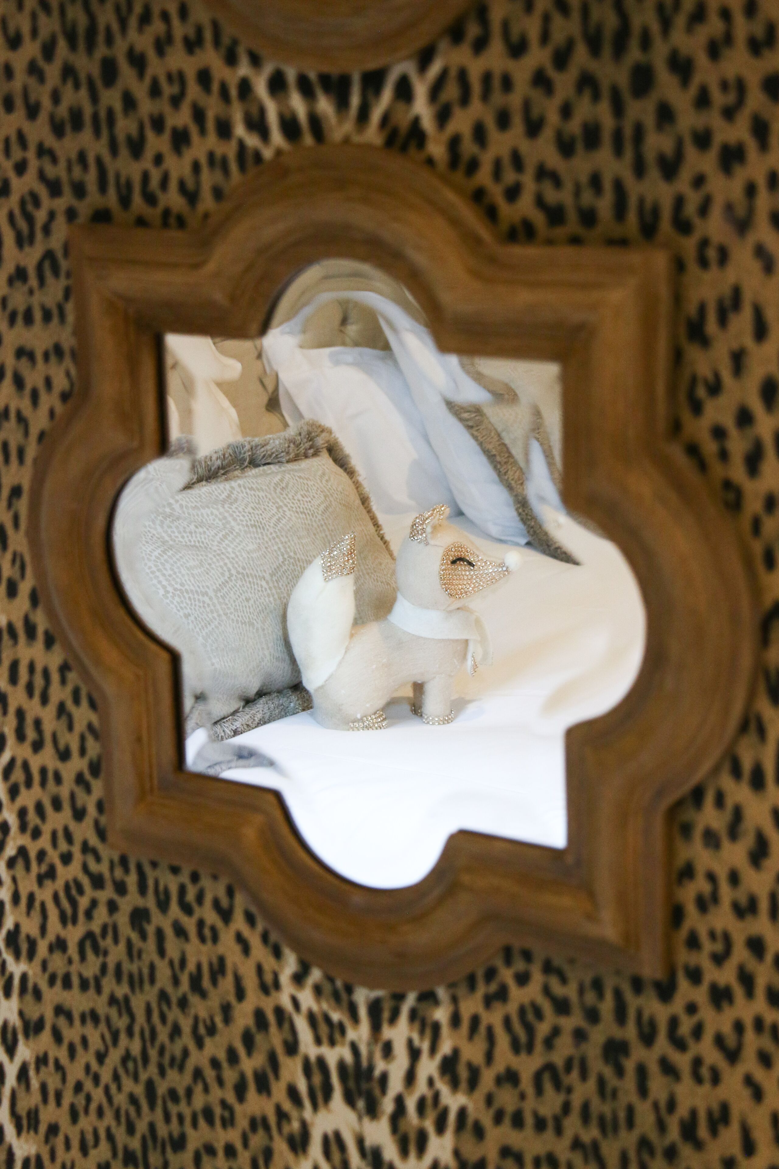 A mirror with a wooden frame reflecting a bed with white bedding and a decorative pillow.