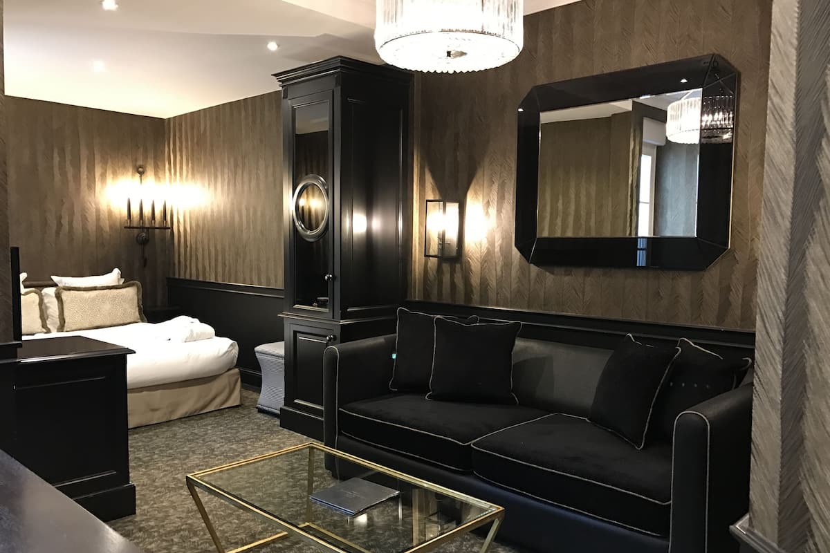 A modern hotel room with a black leather sofa, a glass coffee table, a white bed, and a large mirror.