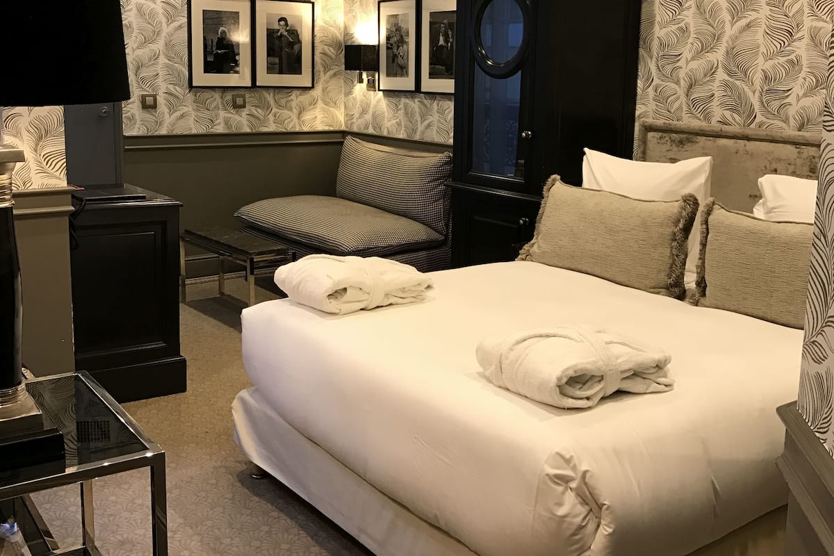 A neatly made bed with two towels, a bedside table with a lamp, a chair, and a wall with framed pictures.