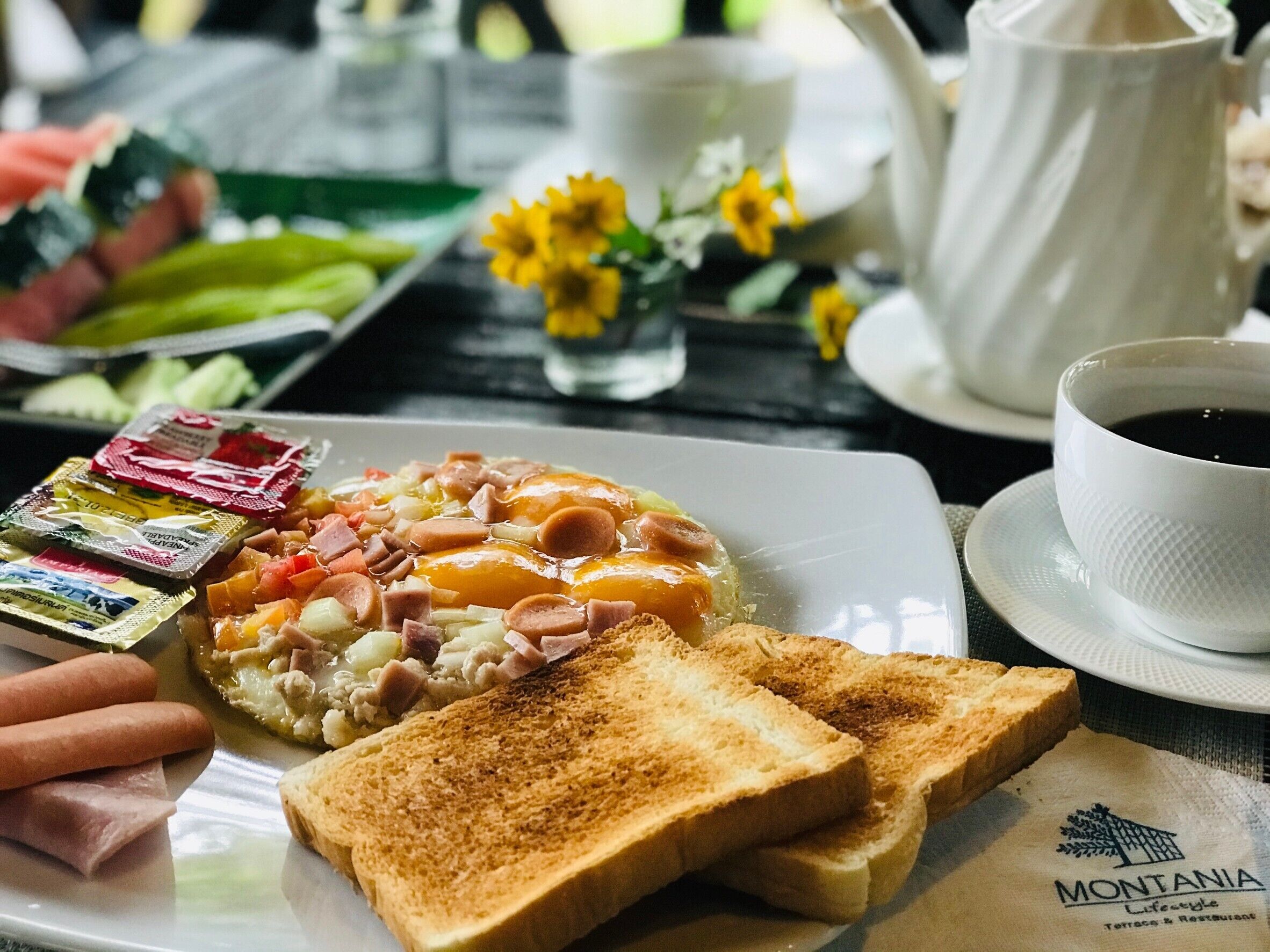 A breakfast plate with eggs, ham, and toast, a cup of coffee, and a teapot on a table.