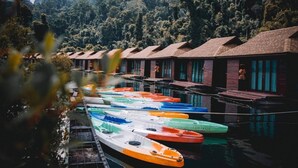 A row of colorful boats docked at a waterfront hotel with wooden buildings and a forested backdrop. - Montania Lifestyle Hotel (Phanom)