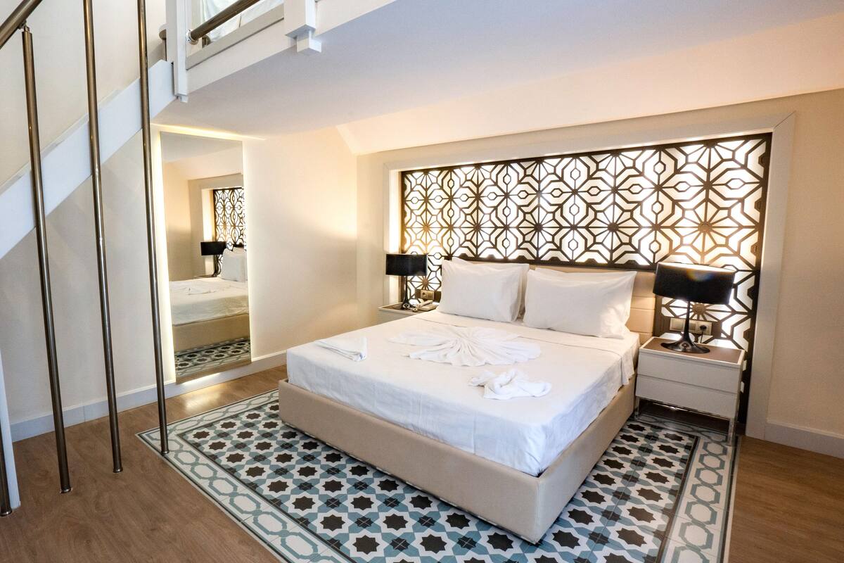 Une chambre moderne avec un grand lit, une tête de lit à motifs, des lampes de chevet et un escalier.