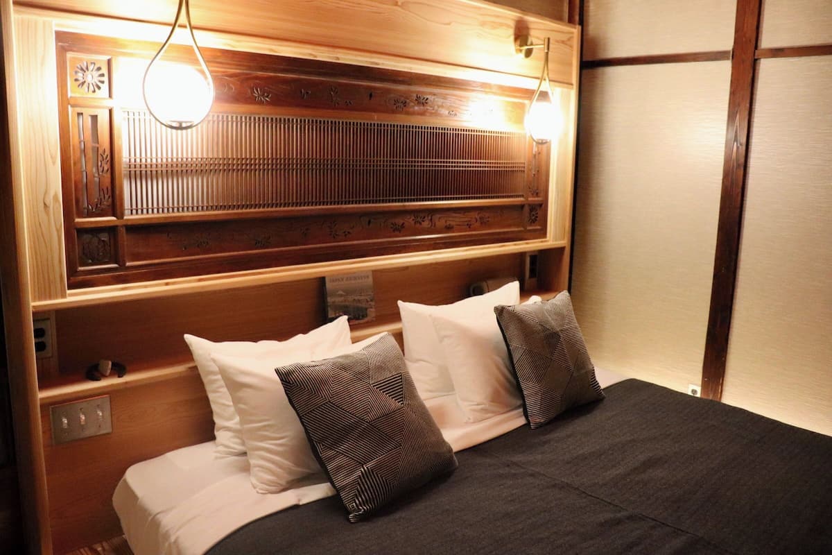 Cama com almofadas brancas e estampadas, colcha escura e cabeceira de madeira com luminária.