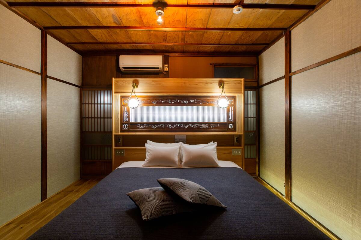 Quarto tradicional japonês com tatames, cama de madeira baixa com colcha azul-escuro, duas almofadas e cabeceira decorativa com luminária.