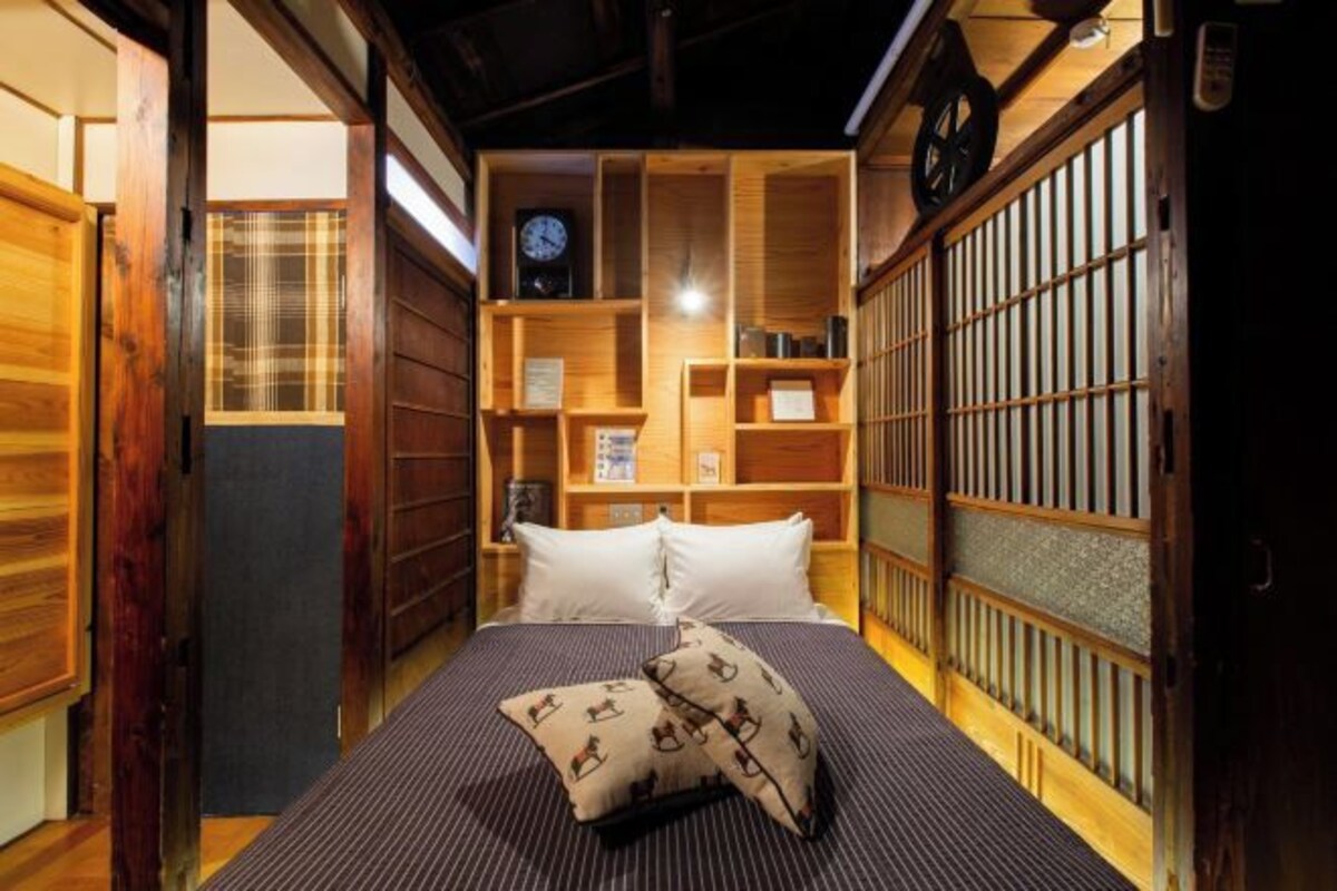 A traditional Japanese-style room with tatami flooring, a bed with a striped bedspread, and wooden shelves with various items.