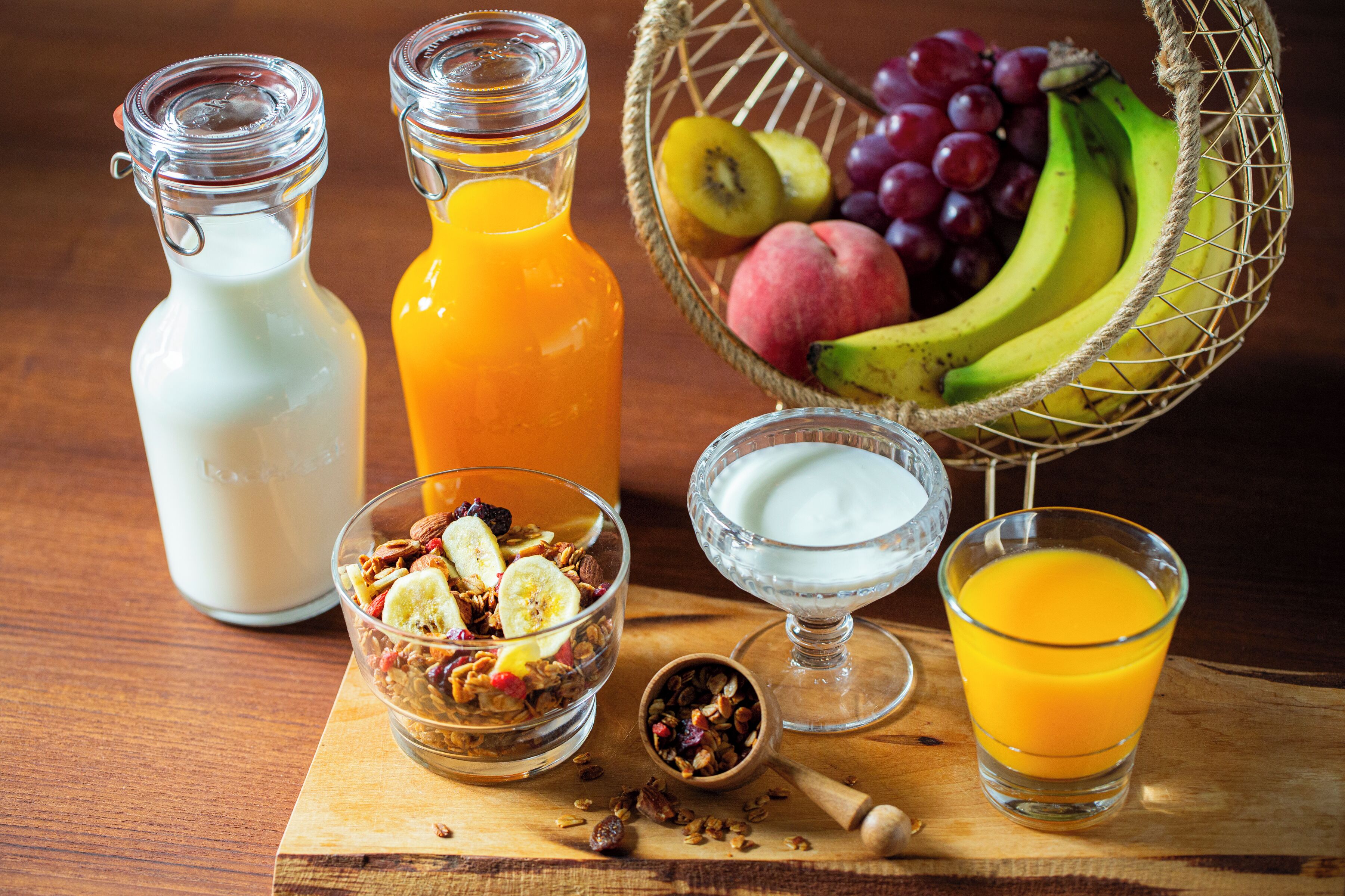 A breakfast setting with a wooden table, two glass bottles, a bowl of granola, a glass of orange juice, a glass of milk, and a basket of fruits.