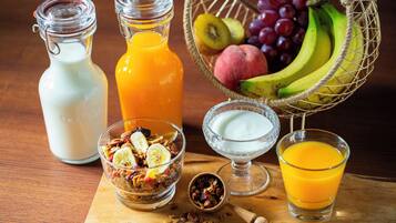 A breakfast setting with a wooden table, two glass bottles, a bowl of granola, a glass of orange juice, a glass of milk, and a basket of fruits.