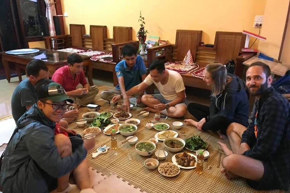 Een groep mensen zit op een mat rond een tafel met verschillende gerechten, in een kamer met houten meubels en een raam.