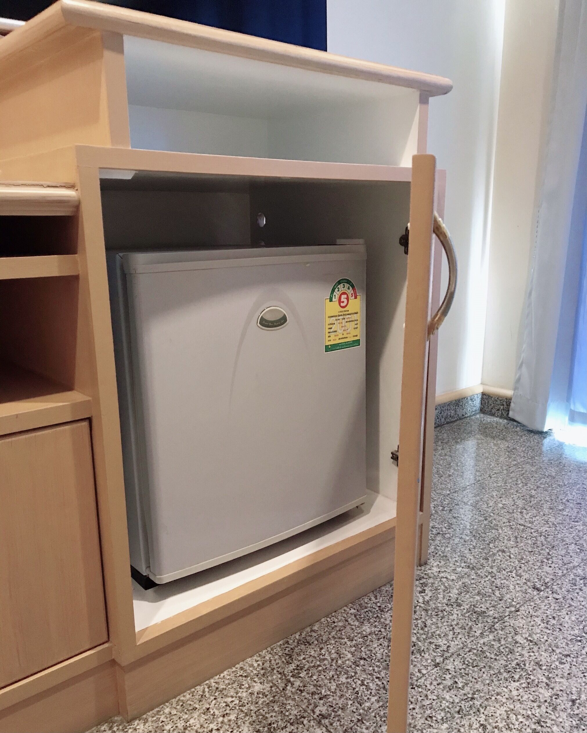 A compact, white refrigerator with a sticker indicating a 5-star rating, housed in a wooden cabinet with a handle, situated in a room with a tiled floor.