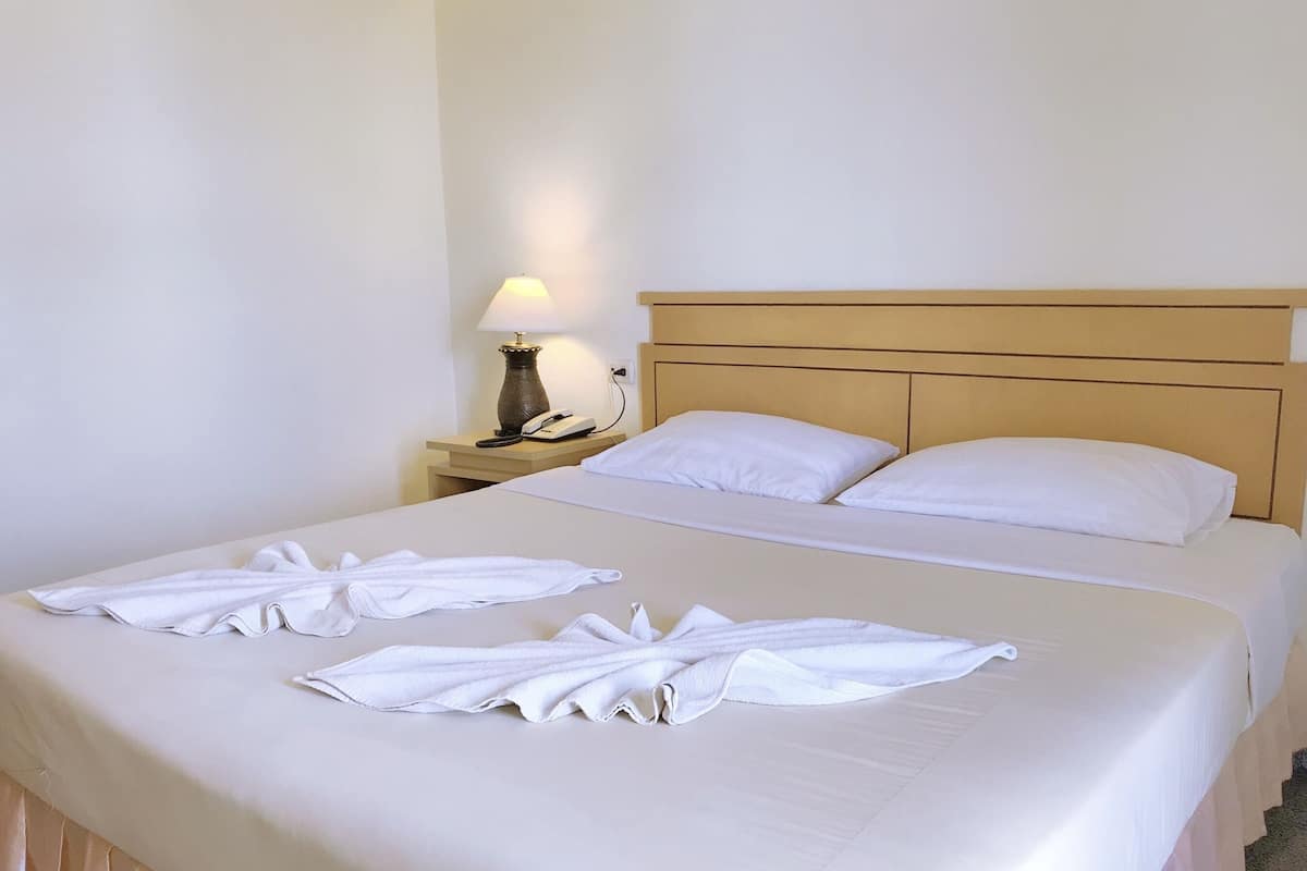 A hotel room with a large bed, two pillows, a bedside table with a lamp, and two towels arranged on the bed.