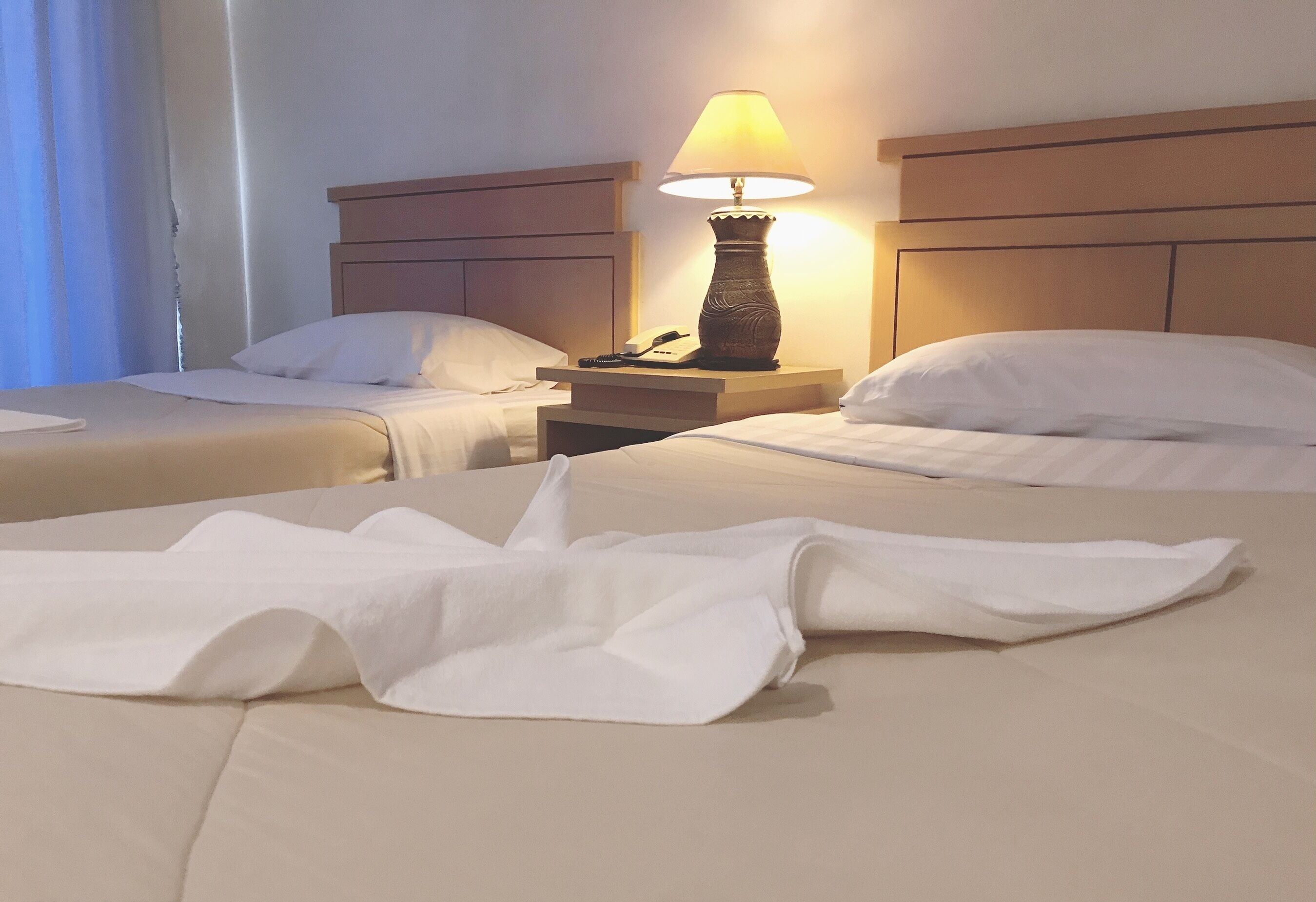 A hotel room with two beds, a nightstand with a lamp, and a window with curtains.