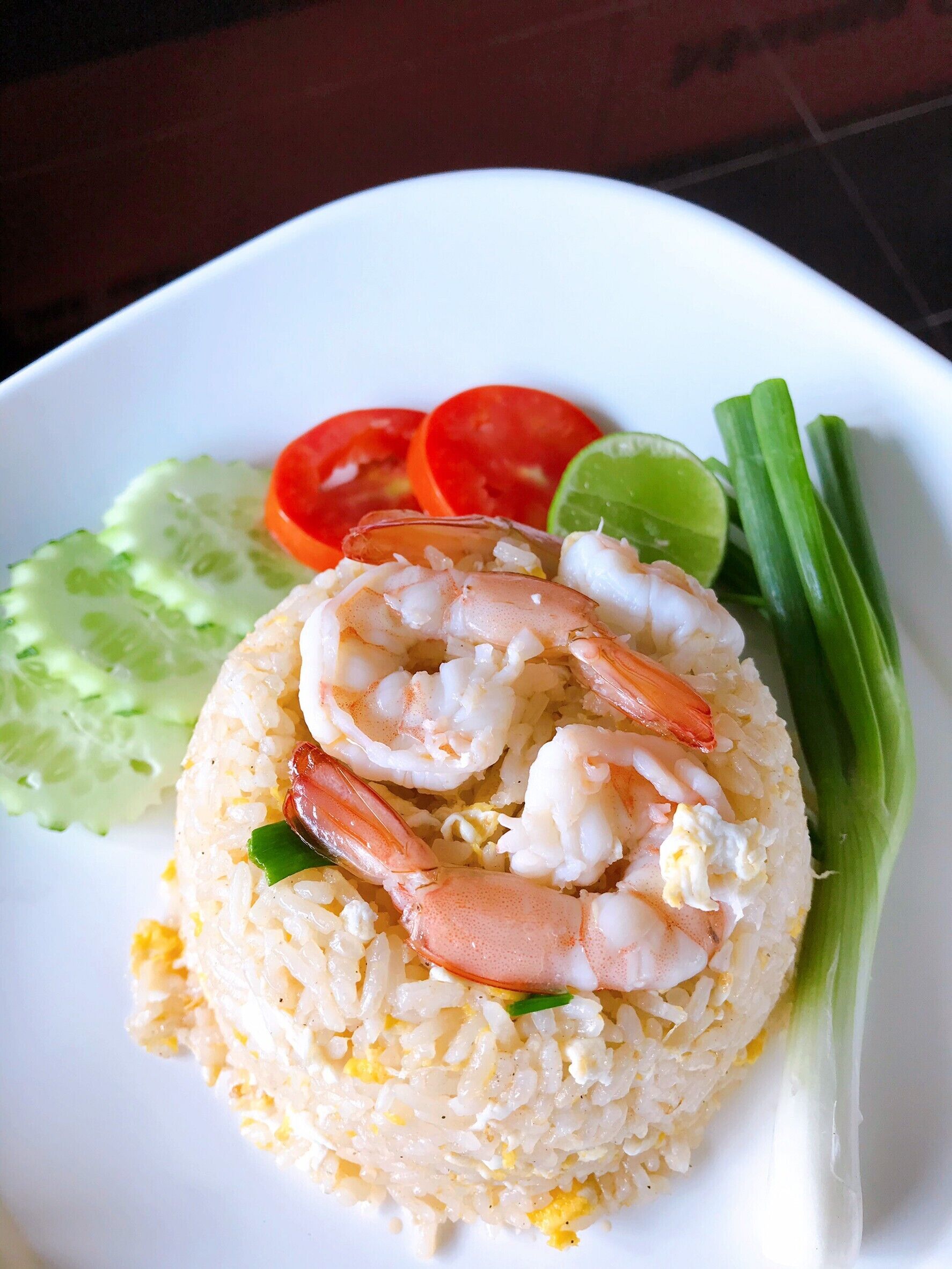 A plate of fried rice topped with shrimp, accompanied by cucumber slices, tomato slices, green onions, and a lime wedge.