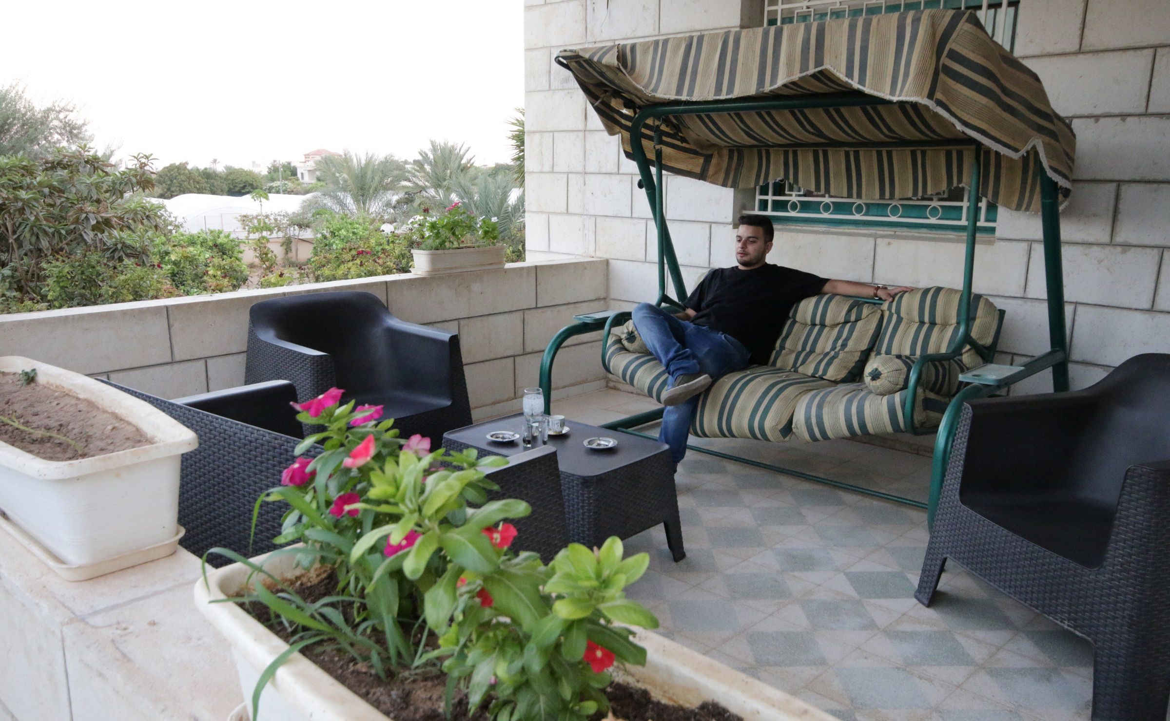 Un patio con mobiliario exterior, incluyendo una silla colgante y dos sillas, rodeado de plantas en macetas y una mesa con un vaso.