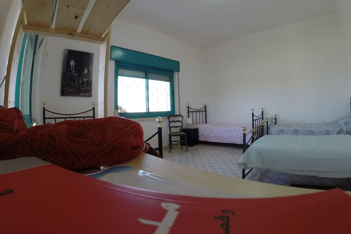 Ein Schlafzimmer mit zwei Betten, einem Schreibtisch und einem Stuhl. Es gibt ein Fenster mit blauer Umrandung und ein Bild an der Wand.